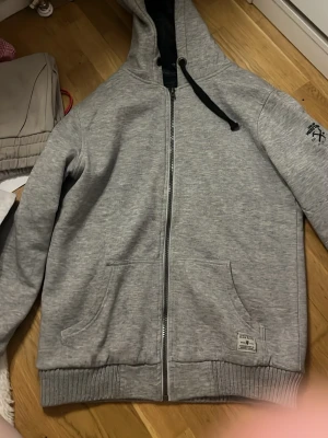 Grå zip hoodie Golden Heritage - Snygg grå zip hoodie från Golden Heritage med svart tryck på ärmen och två stora fickor framtill. Huva med svart insida och dragsko. Tillverkad i mjukt sweatshirtmaterial, perfekt för chill dagar.