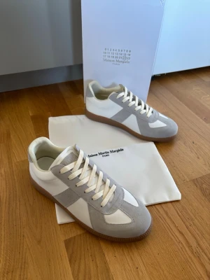 Maison Margiela vita och grå sneakers - Maison Margiela sneakers i vitt och ljusgrått med klassisk låg silhuett. Ovandel i mocka och läder, vita skosnören och brun gummisula. Snygg och stilren design som passar till det mesta. Kommer med dustbag och originalkartong.