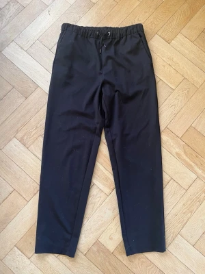 Svarta mjukisbyxor med dragsko - Svarta mjukisbyxor med rak passform och elastisk midja med dragsko. Byxorna har sidfickor och diskreta bakfickor. Perfekta för chill och avslappnade dagar. Materialet är mjukt och bekvämt, vilket gör dem till ett grymt val för dig som vill ha både stil och komfort.