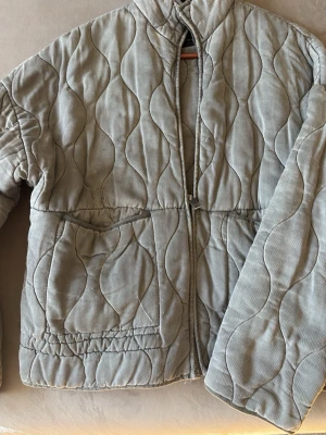 Quiltad grå jacka från Zara - Quiltad grå jacka från Zara med vågigt mönster och dragkedja framtill. Jackan har två fickor och hög krage, perfekt för att hålla värmen under kyliga dagar. Materialet är mjukt och känns bekvämt mot huden.