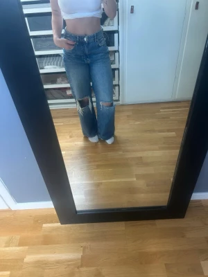 Blåa raka jeans med slitningar - Säljer ett par blåa jeans med raka ben och stora slitningar på knäna. Jeansen har hög midja och klassisk femficksdesign. Perfekta för dig som gillar en avslappnad och trendig look. Materialet är denim. *Större än de ser ut!