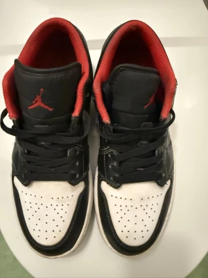 Svarta och vita Air Jordan sneakers - Säljer ett par Air Jordan sneakers med svart och vitt läder, perforerad tå och röda detaljer på insidan samt Jumpman-loggan på tungan. Klassisk låg modell med snörning och cool design som sticker ut. Perfekta för dig som gillar streetstyle och ikoniska sneakers.