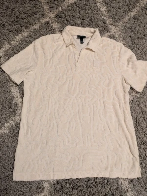 Beige frottéskjorta från H&M x Keith Haring - Säljer en beige skjorta från H&M x Keith Haring med frottématerial och subtilt mönster i tyget. Skjortan har krage och korta ärmar, perfekt för dig som gillar unik design och sköna material.