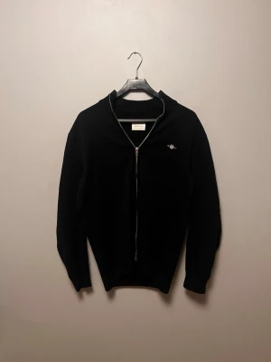 Svart zip-tröja från GANT - Svart tröja från GANT med hel dragkedja och ribbad krage. Diskret broderad logotyp på bröstet. Perfekt för lager-på-lager och enkel att matcha med jeans eller chinos. Materialet är stickat och ger en klassisk look.