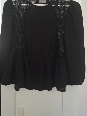 Svart blus med spetsdetaljer - Snygg svart blus från Vera Moda med transparenta spetsdetaljer upptill och längs sidorna. Blusen har lång ärm, rund halsringning och volang nedtill för en feminin look. Perfekt att styla med jeans eller kjol.