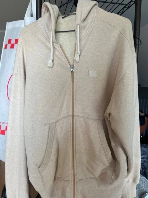 Beige zip-hoodie från Acne Studios - Säljer en beige hoodie från Acne Studios med dragkedja, stor huva och snören. Hoodien har två stora fickor framtill och diskret logopatch på bröstet. Super skön. Passformen är lite oversize.