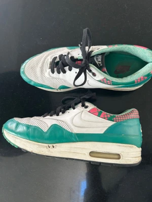 Nike Air Max 1 retro sneakers - Nike Air Max 1 sneakers med i läder, vita och gröna med rosa detaljer och svart snörning. Insidan har grönt foder. Klassisk Air Max-sula med synlig luftbubbla. Perfekt för dig som gillar retrostil och streetwear.