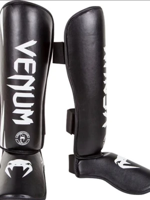 Venum svarta benskydd - Säljer ett par svarta benskydd från Venum med vit logga och detaljer. Benskydden är tillverkade i syntetiskt läder och har kardborreband för enkel justering. Perfekta för kampsport och träning där extra skydd behövs.