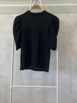 Svart glittrig topp med puffärm - Snygg svart topp med glittrigt tyg och puffiga axlar. Toppen har en rak passform och trekvartsärm som ger en trendig siluett. Perfekt för dig som vill sticka ut lite extra. Materialet känns mjukt och stretchigt.