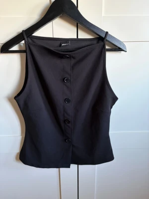 Svart ärmlös linnetopp med knappar - Snygg svart linnetopp från Monki med raka axelband och knappar framtill. Toppen är ärmlös och har en stilren, enkel design som passar perfekt till många olika outfits. Materialet känns mjukt och lätt mot huden.