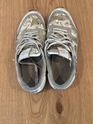 Grå sneakers med stjärnmönster från Atlantic Stars - Snygga sneakers från Atlantic Stars med grå och beige färgtoner, dekorerade med guldiga och vita stjärnor på sidorna. Skorna har klassiska snörningar och meshdetaljer vid hälen för extra komfort. Perfekt för dig som vill sticka ut med coola detaljer.