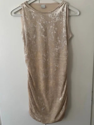 Beige sammet klänning omlott - Säljer en beige kort klänning i mjuk sammet med omlottdetalj på sidan. Klänningen har en lätt glansig yta, perfekt för dig som gillar en stilren men ändå unik look.