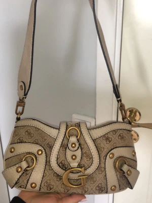 Beige handväska från Guess - Cool beige handväska från Guess med mönstrad canvas och detaljer i ljus läderlook. Väskan har guldiga metalldetaljer, bred axelrem och ett stort G på framsidan. Perfekt för dig som gillar statement-accessoarer och snygga detaljer. Den är även äkta och köpt från Åhléns.