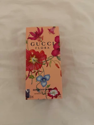 Gucci Flora Gorgeous Gardenia parfym - Gucci Flora Gorgeous Gardenia parfym i en snygg rosa kartong dekorerad med färgglada blommor i rött, blått och vitt. Förpackningen har en lyxig och ungdomlig känsla, perfekt för dig som gillar exklusiva dofter och vacker design.