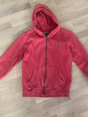 Röd hoodie från Lyle & Scott - Röd hoodie från Lyle & Scott med dragkedja och huva. Storlek 14-15 år. Det står inte centimeter i tröjan men den passar bra för 158/164 cm bedömer jag.  Märke: Lyle & Scott  Obs!. En liten slitning på vardera ärmslut enligt bilder