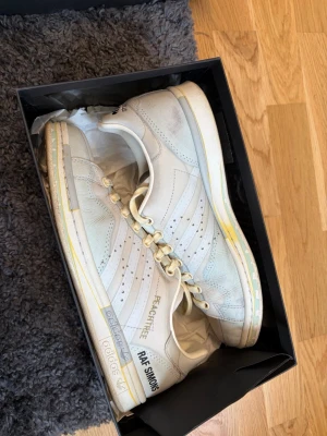 Adidas Raf Simons Stan Smith vita sneakers - Unika vita sneakers från Adidas x Raf Simons, Stan Smith-modell. Skorna har klassiska perforerade ränder, gulddetaljer och text på sidan. Tillverkade i läder med lågt skaft och ikonisk Adidas-logga på hälen. Perfekta för dig som gillar stilrena och trendiga sneakers.