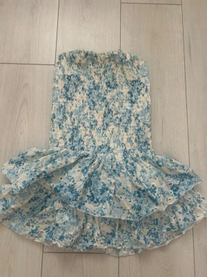 LVCILIA Klänning - Söt kort klänning med smockad överdel, volangkjol och inbyggda shorts. Klänningen har t ett blått blommönster på vit botten. Perfekt för varma dagar och har en luftig känsla. Materialet känns lätt och somrigt.