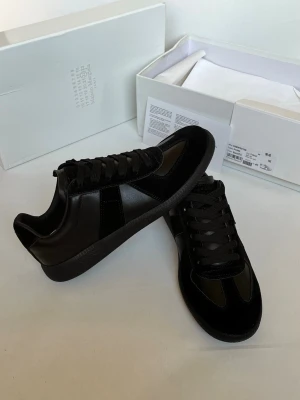 Svarta sneakers från Maison Margiela - Säljer ett par svarta sneakers från Maison Margiela med klassisk låg profil. Skorna har en mix av läder och mocka, svarta skosnören och en svart yttersula. Perfekta för dig som vill ha en stilren och tidlös look.