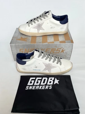 GGDB Superstar vita sneakers - Snygga GGDB Superstar sneakers i vitt läder med grå stjärndetalj på sidan och mörkblå häl. Grå skosnören och beige sula ger en cool kontrast. Klassisk låg modell med tydlig branding och retrovibe. Perfekta för dig som gillar streetstyle och unika detaljer.