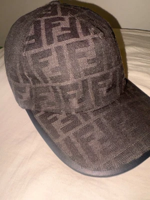 Mörkgrå/svart Fendi keps med FF-mönster - Säljer en mörkgrå keps från Fendi med det ikoniska FF-logomönstret över hela kepsen. Kepsen är tillverkad i ett tygmaterial och har en böjd skärm med en snygg finish. Märkt med Fendi Roma på insidan och tillverkad i Italien. Perfekt för dig som gillar exklusiva accessoarer.