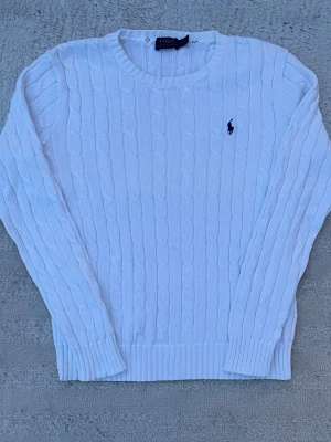 Vit kabelstickad tröja från Polo Ralph Lauren - Säljer en vit kabelstickad tröja från Polo Ralph Lauren med klassisk rund halsringning och broderad logotyp på bröstet. Tröjan har långa ärmar och ribbade muddar vid ärmslut och nederkant. Perfekt för dig som gillar stilrena och tidlösa plagg.