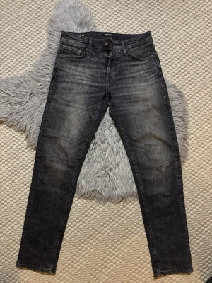 Svarta tapered mike jeans från Jack & Jones - Säljer ett par svarta tapered mike jeans från Jack & Jones med snyggt slitna detaljer och klassisk femficksdesign. Jeansen har knappgylf och är tillverkade i bomullsdenim med lite stretch för extra komfort. Perfekt för dig som gillar en modern och avslappnad look.