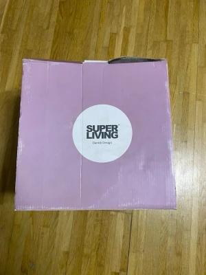 Rosa kartong från Super Living - Säljer en rosa kartong från Super Living med vit cirkel och logotyp på framsidan. Kartongen har dansk design och är tillverkad i papp. Perfekt för förvaring eller som presentlåda.