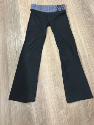 LOVE PINK svarta leggings bootcut - Säljer ett par svarta leggings från Victoria's Secret PINK med grå, ribbad midja och texten 'LOVE PINK' tryckt framtill. Modellen har bootcut-ben och är tillverkad i mjukt, stretchigt material som sitter skönt på kroppen.
