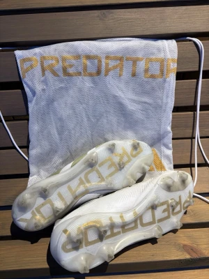 Adidas Predator vita fotbollsskor - Vita Adidas Predator elite fotbollsskor med guldiga detaljer och klassiska ränder. Skorna är knappt använda då de var för små och är därför i mycket gott skick. Skorna är i storlek 45 1/3 men passar 45 och de har gräsdobbar. Påse och box tillkommer! Nypris är 3099kr