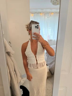 Vit ärmlös linnetopp med knyt - Säljer en vit, ärmlös linnetopp med halterneck och öppen rygg. Toppen har smockade detaljer, knyt i nacken och ryggen samt en lätt volangkant nedtill. Perfekt för sommaren och ger en fräsch, ljus look.