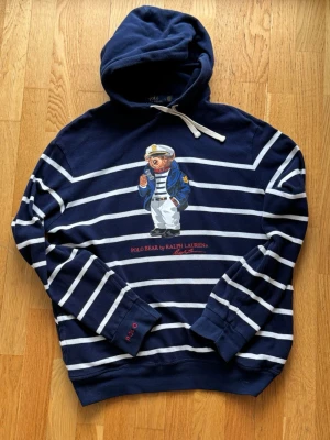 Ralph Lauren Hoodie - En unik hoodie från Polo Ralph Lauren med den ikoniska Polo Bear i sjömansstil. Modellen har en marininspirerad design med mörkblå bas och vita ränder. Tillverkad i lätt bomull, vilket gör den perfekt för varmare väder.