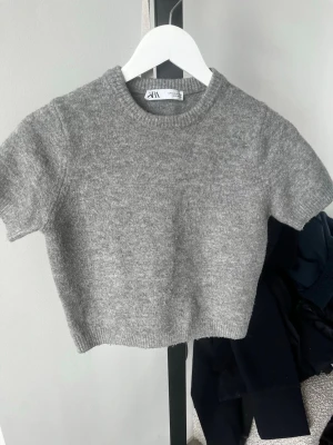 Grå stickad kortärmad tröja Zara - Mysig grå stickad tröja från Zara med korta ärmar och rund hals. Tröjan har en enkel, rak passform och ribbade detaljer vid hals, ärmslut och nederkant. Perfekt att styla med jeans eller kjol för en chill look.