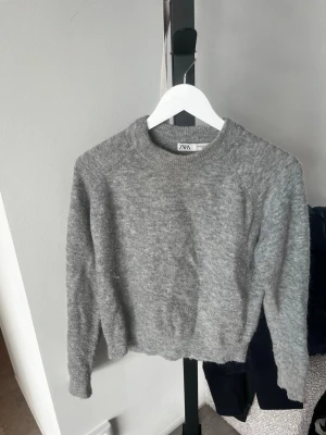 Grå stickad tröja från Zara - Mysig grå stickad tröja från Zara med rund halsringning och långa ärmar. Tröjan har en enkel, klassisk design och är gjord i ett mjukt, fluffigt material som känns skönt mot huden. Perfekt för kyliga dagar och enkel att matcha med olika outfits.