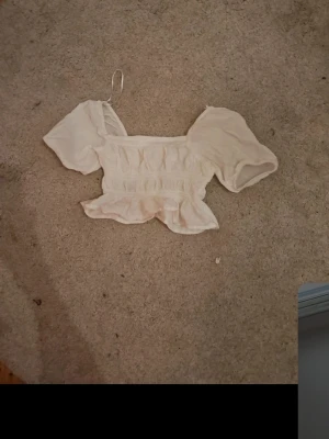 Vit croppad blus med puffärm - Söt vit croppad blus med puffiga korta ärmar och smockad midja. Toppen har volangkant nedtill och en luftig, romantisk look. Perfekt för dig som gillar trendiga och feminina plagg.