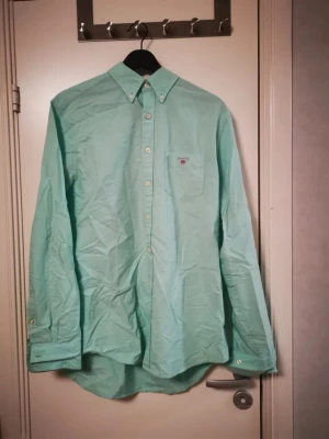 Turkos skjorta från Gant - Snygg turkos skjorta från Gant med klassisk button-down krage och bröstficka med liten broderad logga. Skjortan har långa ärmar och knappar framtill. Perfekt för en stilren och avslappnad look.