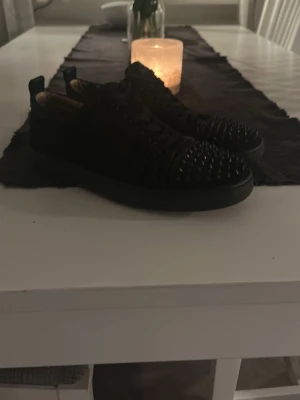 Svarta sneakers med nitar från Louboutin - Säljer ett par svarta sneakers från Christian Louboutin med coola nitar på tån och klassisk röd sula. Skorna har lågt skaft och är tillverkade i läder med snörning framtill. Perfekta för dig som vill sticka ut med din stil.
