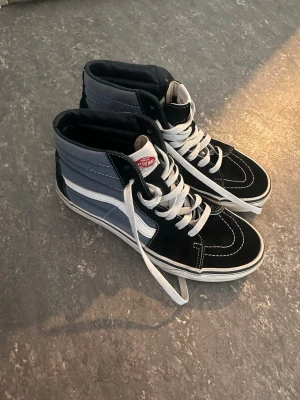 Vans Sk8-Hi svart och blå sneakers - Säljer ett par klassiska Vans Sk8-Hi sneakers i svart och blått med vit sidostripes och vita snören. Skorna har hög modell och är tillverkade i canvas och mocka. Inga tecken på slitningar osv helt som nya. Gör rent skorna innan jag skickar iväg de.  