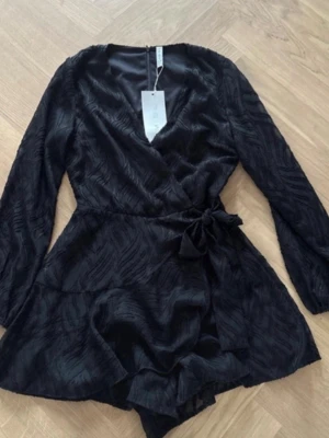 Svart omlottjumpsuit med struktur - Snygg svart jumpsuit med omlottmodell och knytband i midjan. Långa ärmar och lätt glansigt tyg med strukturmönster ger en cool look. Perfekt för dig som gillar trendiga och bekväma plagg. Kort modell med volangdetaljer nertill.