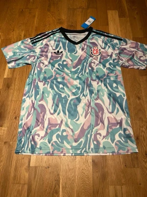 Adidas camo sporttröja pastell - Säljer en Adidas sporttröja med pastellfärgat camomönster i lila, turkos och vitt. Tröjan har svarta detaljer vid kragen och axlarna samt Adidas-logga och badge på bröstet. Tillverkad i lätt och ventilerande material, perfekt för träning.