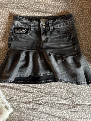 Grå jeanskjol med volangkant - Säljer en grå jeanskjol med volangkant och dubbla knappar i midjan. Kjolen har klassiska jeansfickor fram och bak samt en snygg, lite tvättad look. Perfekt för dig som gillar en edgy men söt stil. Och det bästa är att kjolen har inbyggda   shorts!💘