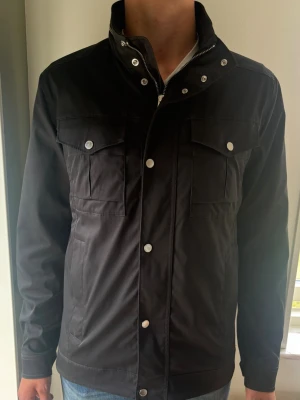 J.Lindeberg Bailey Field Jacket (Vår/Sommar/höst Jacka) - Svart J.Lindeberg Bailey Jacka i storlek L. En exklusiv field jacket riktigt snygga detaljer och en JL logga på ärmen. Perfekt för alla väder och en otroligt snygg jacka.  📏 Storlek L, Regular fit  🧵 96% Polyester 4% Elestan ✅ B Grade. Använd men i mycket gott skick.  ✨ Logopräglade metallknappar, fyrficksdesign💰 Nypris: Ca 3500 kr  ‼️ Skickas snabbt ‼️