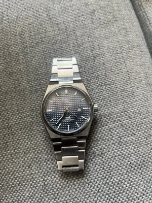 Tissot PRX blå klocka i stål - Säljer en Tissot PRX klocka med blå urtavla och datumvisning. Klockan har ett klassiskt och stilrent armband i rostfritt stål och en rund boett. Urtavlan har ett rutigt mönster och silverfärgade detaljer. Perfekt för dig som gillar snygga accessoarer.