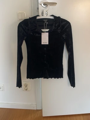 Svart sammetskofta från Monki - Supersnygg svart sammetskofta från Monki med lång ärm och vågig krage. Koftan har knäppning framtill och en glansig finish som ger en lyxig känsla. Perfekt för dig som vill ha något extra till din outfit.