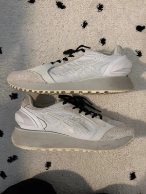 Vita Onitsuka Tiger sneakers - Snygga vita sneakers från Onitsuka Tiger med svart snörning och grå detaljer. Skorna har en chunky sula och är tillverkade i mesh och mocka för en modern look. Perfekta för dig som gillar stilrena och trendiga sneakers. Köpta i Tokyo 2024 men sparsamt använda. Små i storlek, passar 39,5-40.