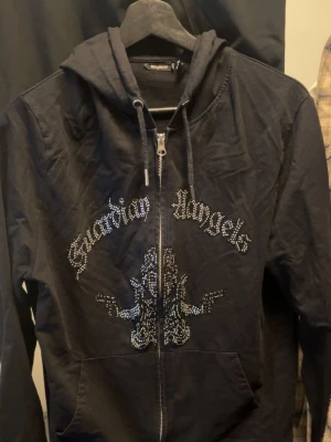 Svart hoodie från Blinglucid storlek M - Säljer en svart hoodie från Blinglucid med dragkedja och huva. På bröstet står det 'Guardian Angels' i glittrande stenar och ett stort bling-motiv under texten. Hoodien har fickor framtill och snören i huvan. Perfekt för dig som gillar streetwear och statement-plagg. Storlek M 