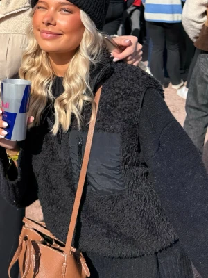 Svart fleeceväst med dragkedja - Säljer en svart fleeceväst med dragkedja och quiltade detaljer på bröstet. Västen har en croppad passform och är perfekt att slänga över en hoodie eller stickad tröja. Materialet är mjukt och fluffigt, vilket gör den både bekväm och snygg.