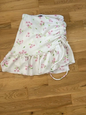 Vit kjol  - Söt vit kortkjol med rosa blommor och gröna blad, snörning på sidan och volang längst ner. Kjolen är gjord i ett lätt bomullsliknande material och har en omlott-design som ger en trendig och feminin look.