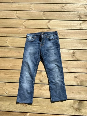 Blå jeans från Nudie Jeans - Säljer ett par klassiska blå jeans från Nudie Jeans med raka ben och snygga slitningar. Jeansen har fem fickor, orangea sömmar och läderpatch bak. Perfekt för dig som gillar en avslappnad och tidlös look.