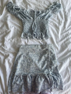 Blått set med volanger från Gina Tricot - Supersött tvådelat set från Gina Tricot i ljusblått med småblommigt mönster. Toppen har off-shoulder, smock och volangkant, kjolen har volang nertill och knytband i sidan. Perfekt för en trendig och feminin look!I