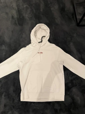 Hugo boss - Vit hoodie med röd logga - Snygg vit Hugo boss hoodie med huva, samt en liten röd logga på bröstet. Klassisk känguruficka framtill och ribbade muddar vid ärmar och nederkant. Perfekt för en clean och stilren look. Den är använd kanske 3-4 gånger, köptes för 2 år sen typ och har bara legat i garderoben då jag tycker den inte passar just min stil nypris när jag köpte var 3000 kr men såg den för 2500 kr idag 